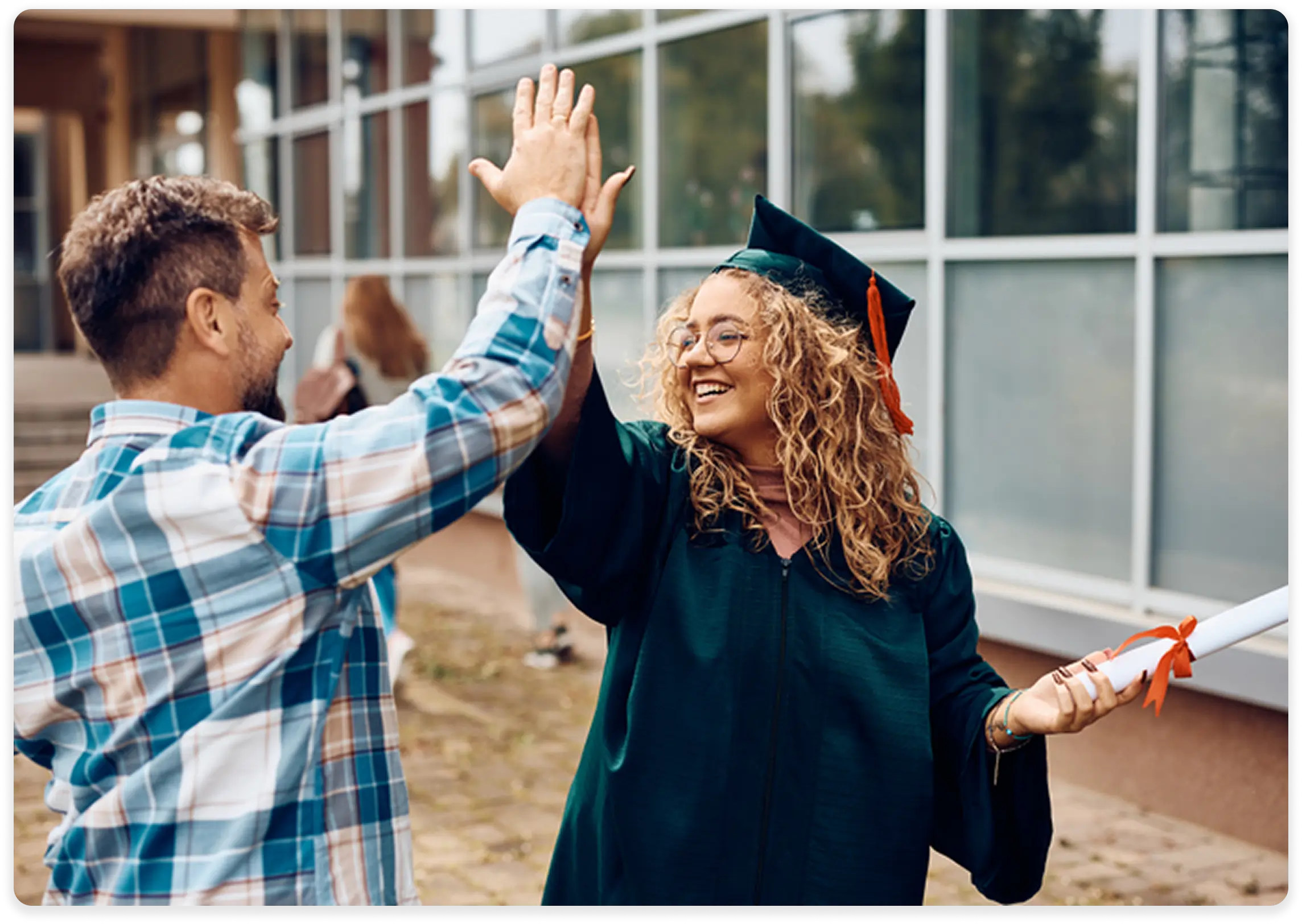 A joyful graduate in a dark green cap and gown is smiling and giving a high-five to a man in a plaid shirt. The graduate is holding a rolled-up diploma tied with a red ribbon. They are standing outside a modern building with large windows, celebrating the achievement.