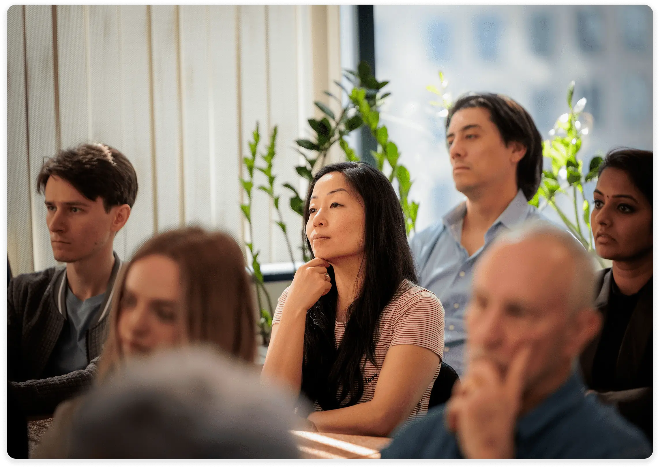 A group of people seated indoors attentively listening to a speaker or presentation. The individuals appear focused and thoughtful, with some resting their chins on their hands. The background features tall windows with vertical blinds and green potted plants, letting in natural daylight. The mood is serious and reflective.