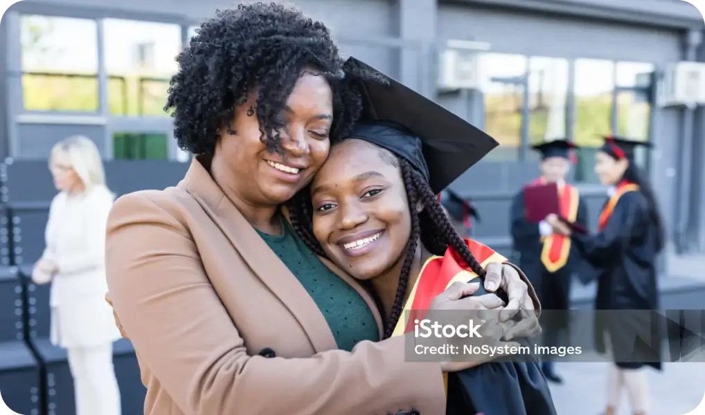 Graduation celebration: A smiling graduate in cap and gown with a red sash embraces a proud companion, surrounded by fellow graduates at an outdoor venue