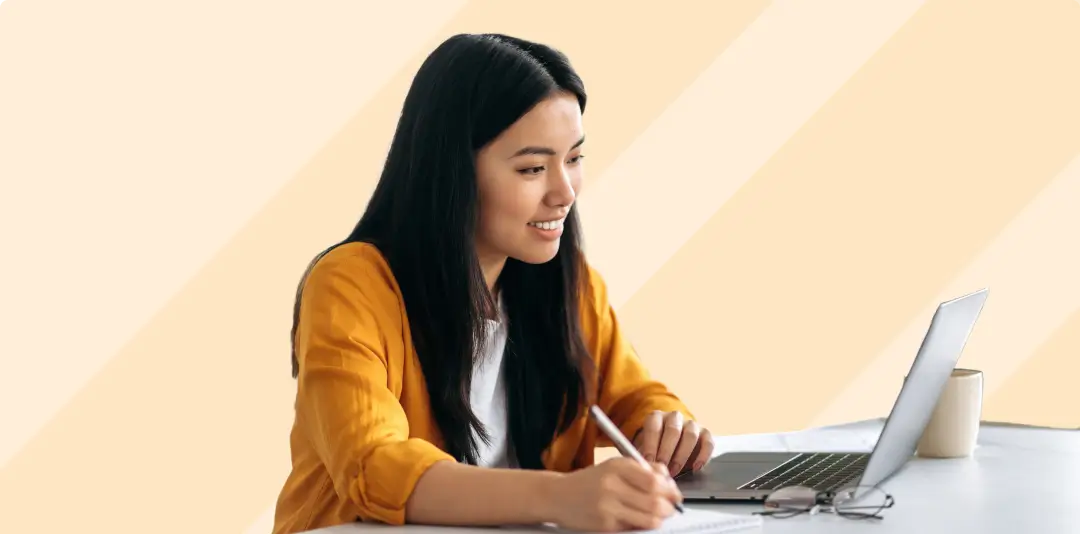 Female professional looking at laptop screen and taking notes, with glasses and a mug on the table