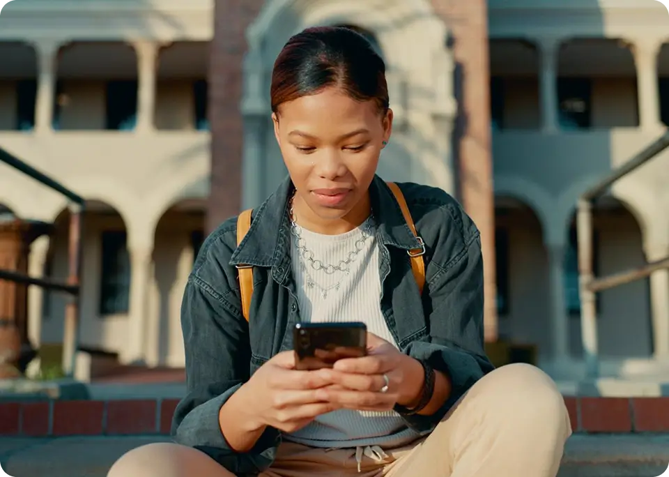 A young female student sitting on the stairs outside a university building and looking down at her smartphone.