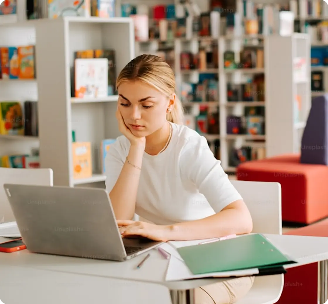 A young blonde female student concentrating and studying on a laptop at a library desk with notebooks and papers.