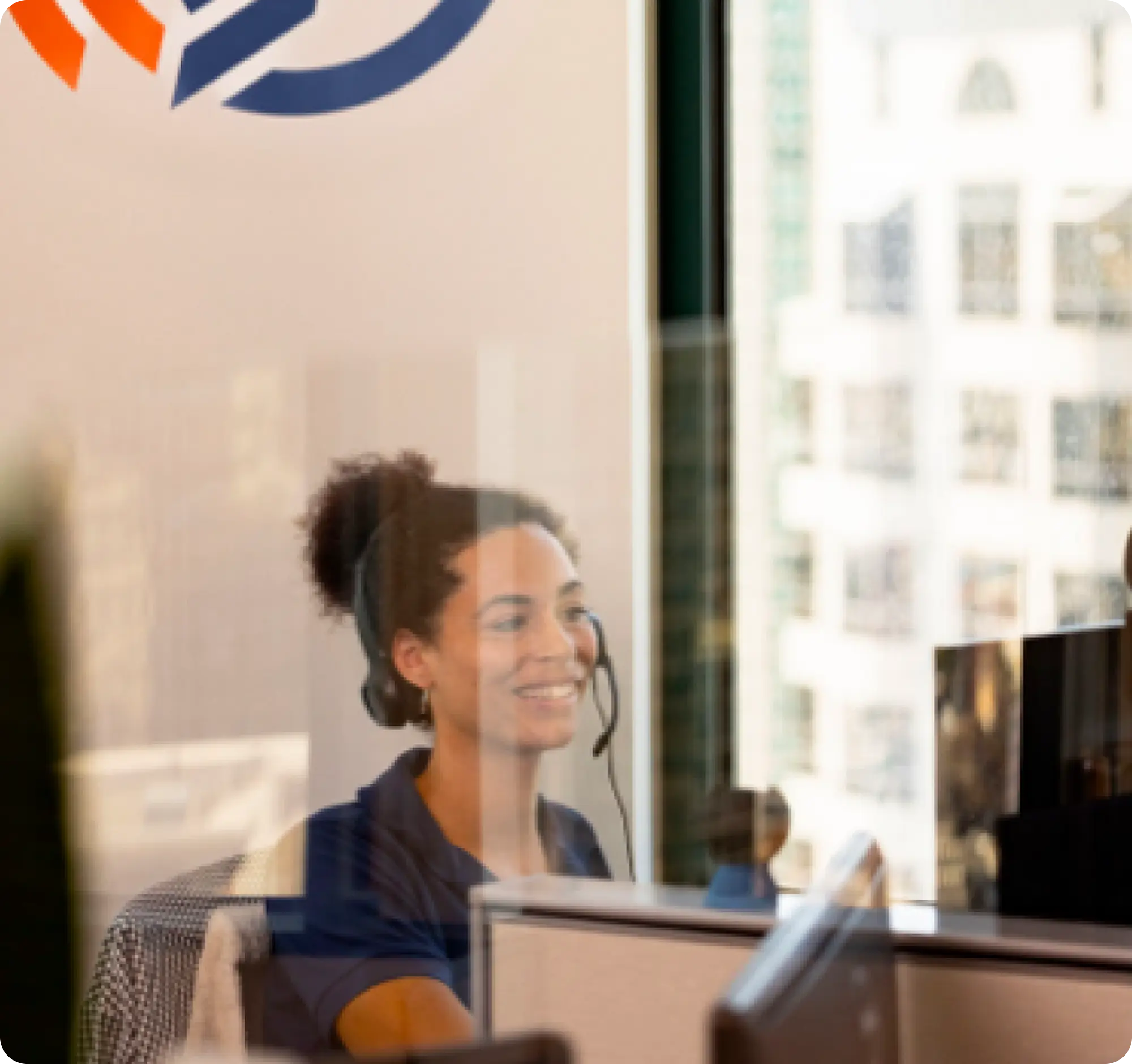 A woman with curly hair, wearing a headset and a blue shirt, smiles while sitting at a desk in an office. She is working in front of a computer, behind a pane of glass. The office building windows reveal a cityscape.