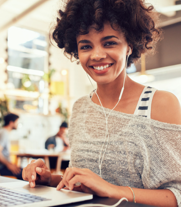 A woman wearing wired headphones, smiling and typing on a laptop