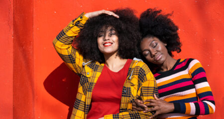 Two women lean on each other, smiling. They stand in front of a red backdrop.