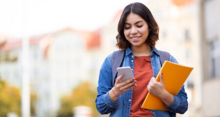 smiling college student looks at her phone while holding a textbook