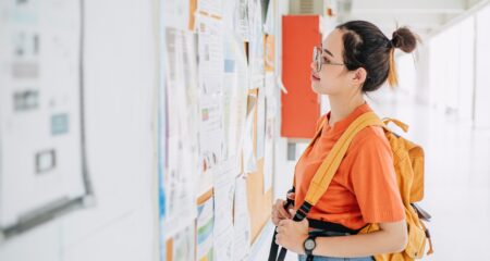 female college student with orange backpack looks at job bulletin board
