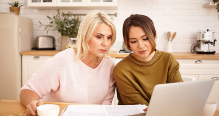 serious mom and teen daughter sit at kitchen table with laptop to correct FAFSA errors