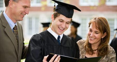 teenage boy in cap and gown looks at high school diploma with parents