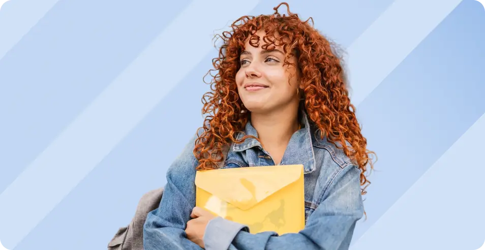 A smiling young woman with curly red hair wearing a denim jacket and holding a yellow plastic folder